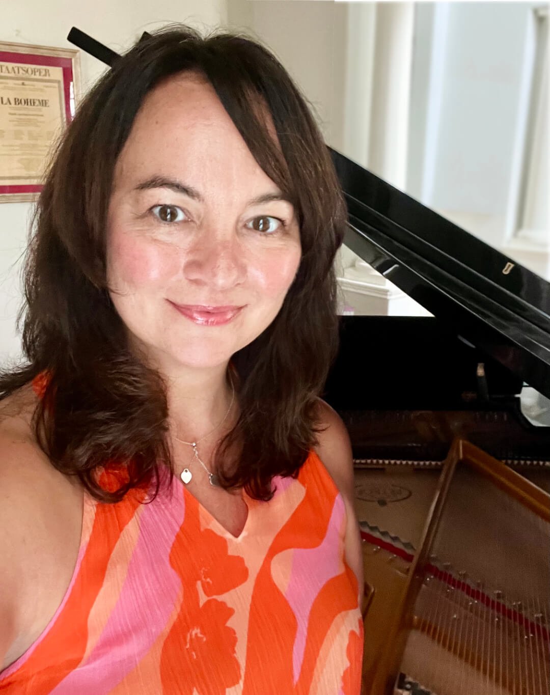 Head and shoulder portrait of a softly smiling woman with shoulder-length brown hair wearing a sleeveless orange and pink patterned top, with a piano in the background