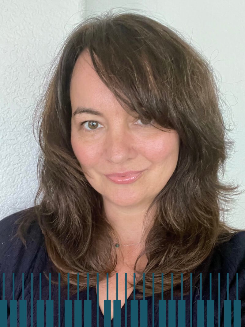 Head and shoulder portrait of a softly smiling woman with shoulder-length brown hair wearing a balck top leaning against a white wall