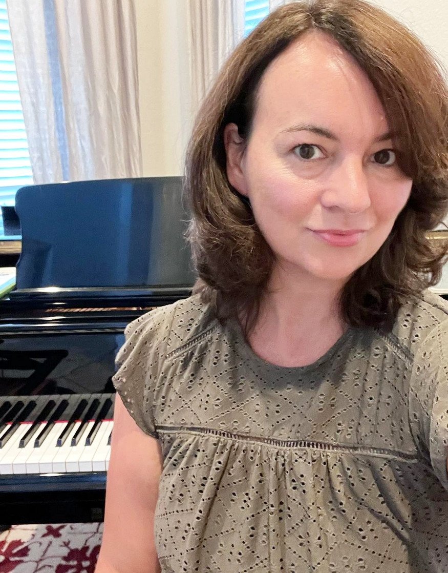 Head and shoulder prortrait of a softly smiling woman with shoulder-length brown hair wearing an olive short sleeve top, with a piano in the background