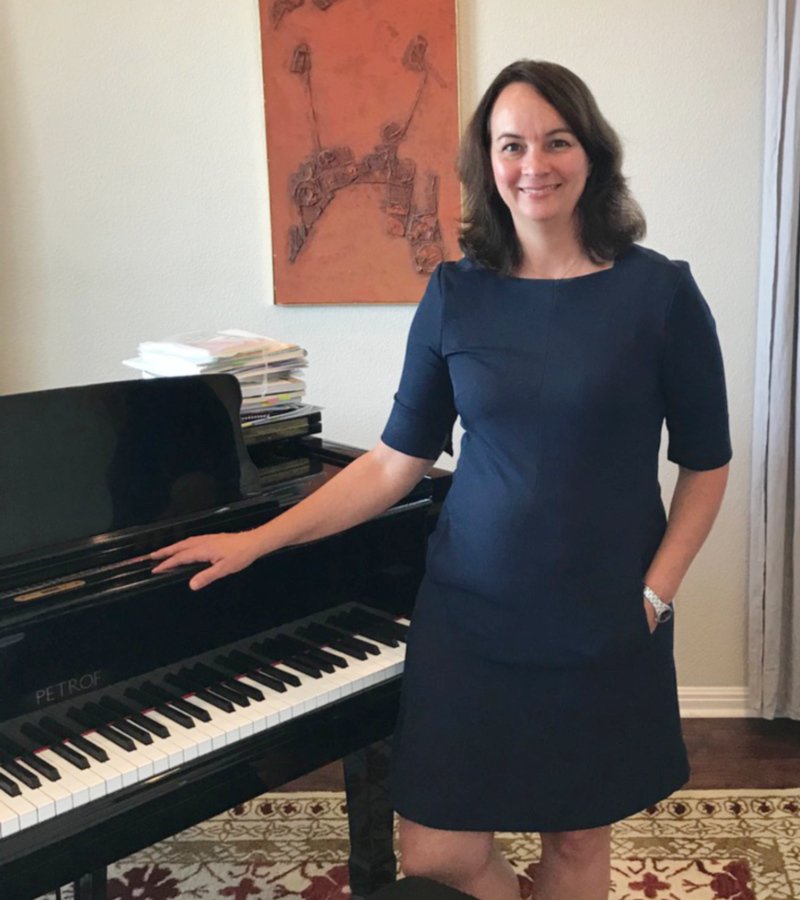 Happily smiling woman with brown hair wearing a dark blue short sleeve dress, standing beside a black piano