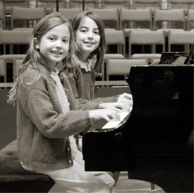 Two young girls smiling at the camera playing the piano together