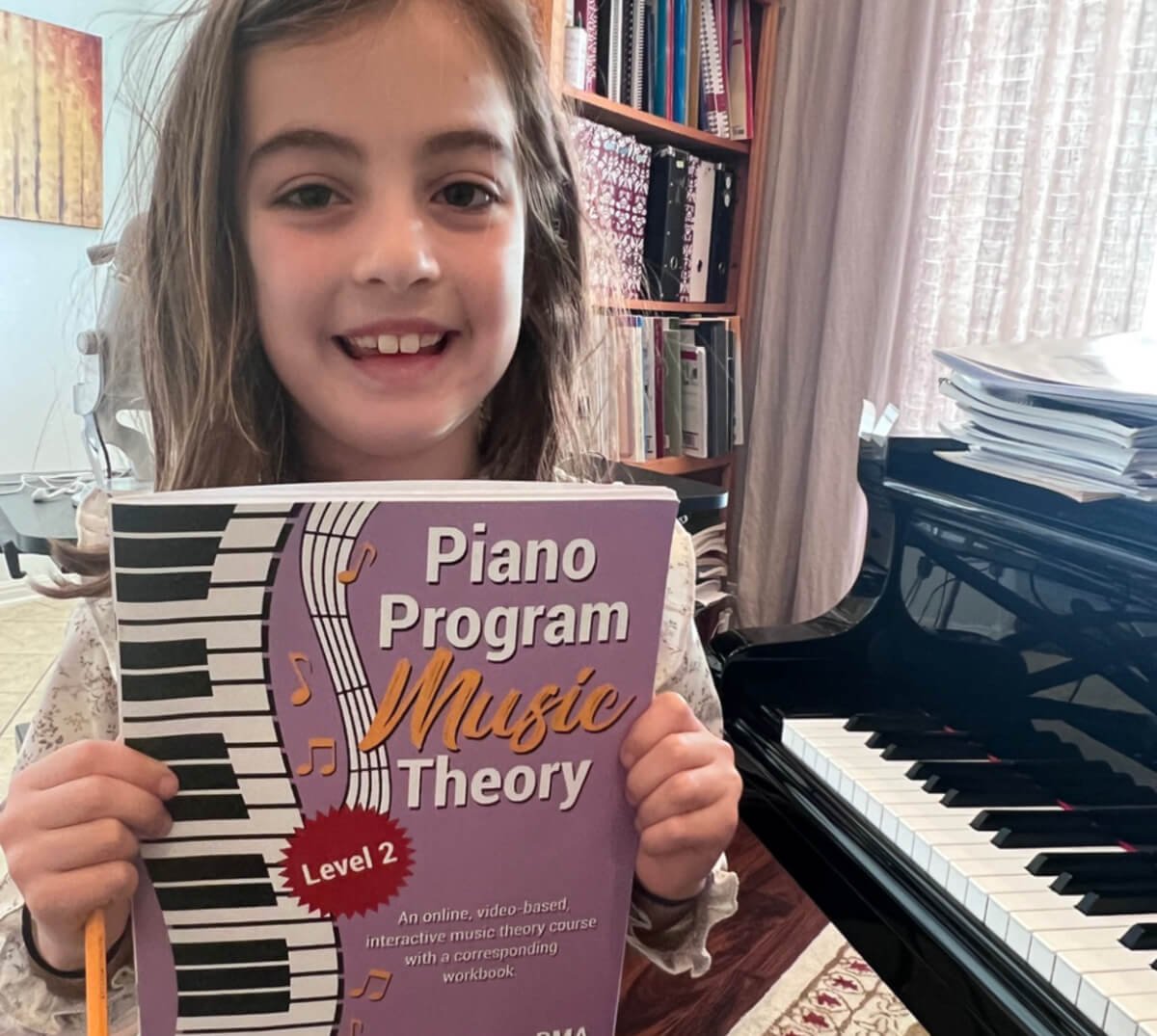 Happily smiling young Caucasian girl with brown shoulder length hair sitting next to her piano holding up a lilac Piano Program Music theory Book